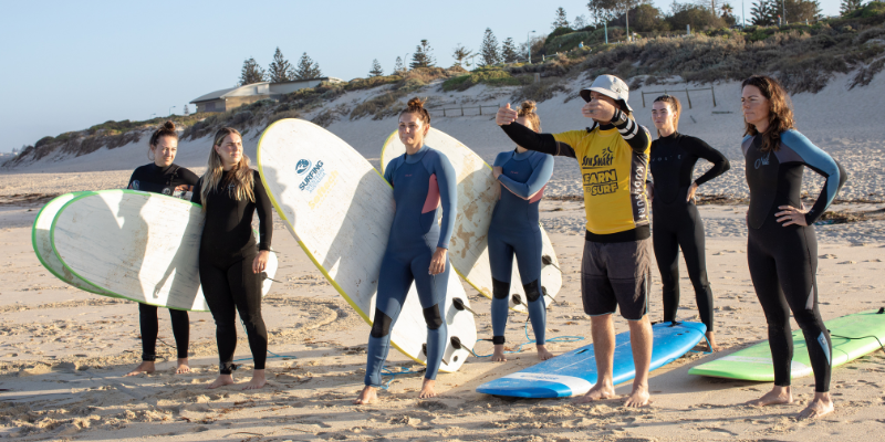 alt= A group of people holding surfboards on a beach in Western Australia, listening to a Surfing WA surfing instructor gesturing with his hand.