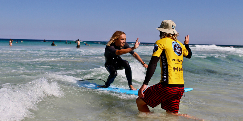 alt= A Surfing WA surf coach hi-fives a student on a surfboard in Western Australia