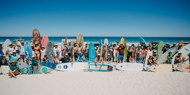 alt= A diverse group of surfers holding surfboards stands on a sanon  sandy western australian beach