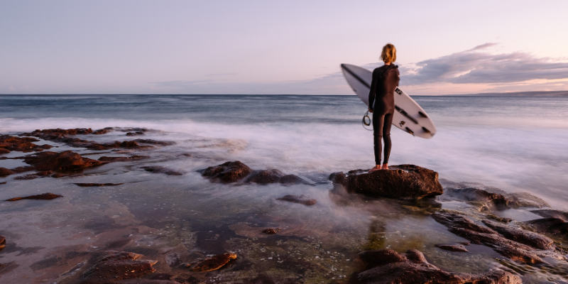 alt= a surfer stands on a rocky shore in Western Australia looking at the ocean during sunset, holding a surf board