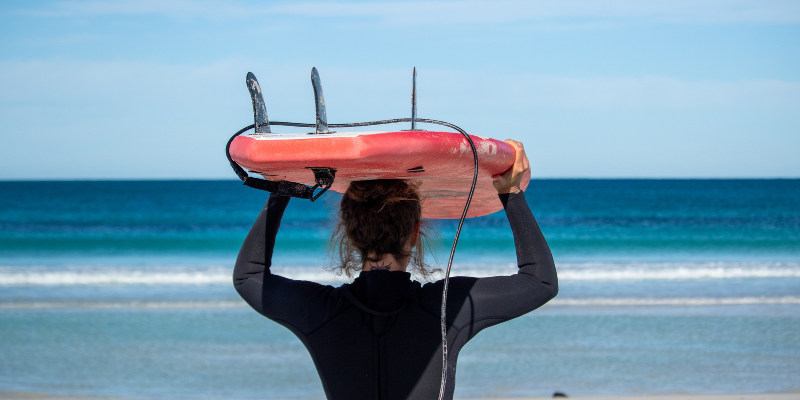 alt= a surfer stands on a rocky shore in Western Australia looking at the ocean during sunset, holding a surf board